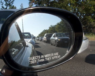 Espejo retrovisor de un auto que muestra otros autos reflejados, se ven más pequeños. En el espejo hay una leyenda en inglés que dice que los objetos pueden estar más cercanos de lo que parece.